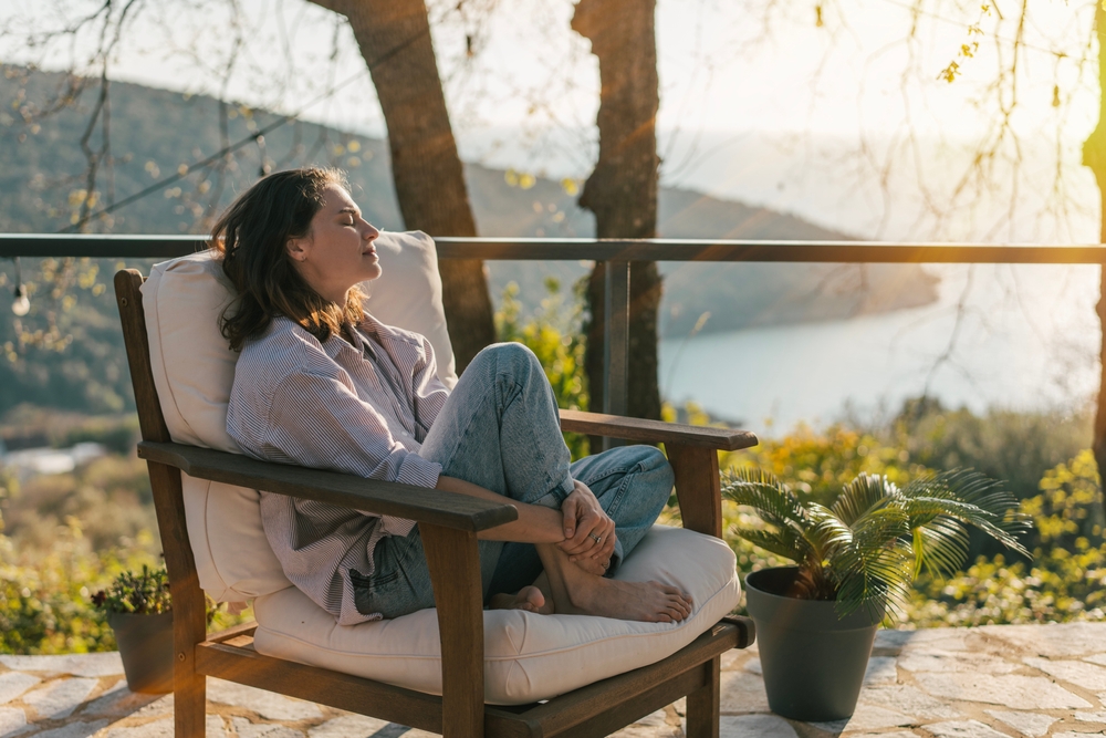 Woman sitting cross-legged in a chair outdoors, quietly reflecting and relaxing, representing self-awareness and compassionate parts work.
